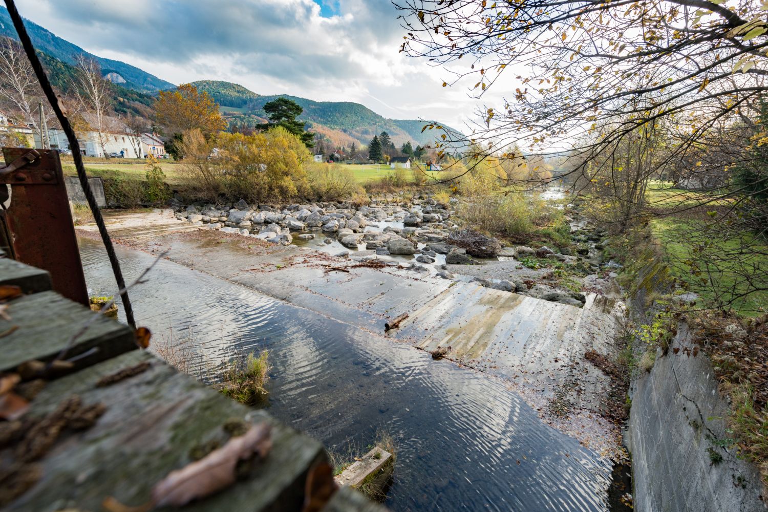 Fluss mit Steinen und Betonwehr in einer bergigen Landschaft mit Bäumen im Herbst