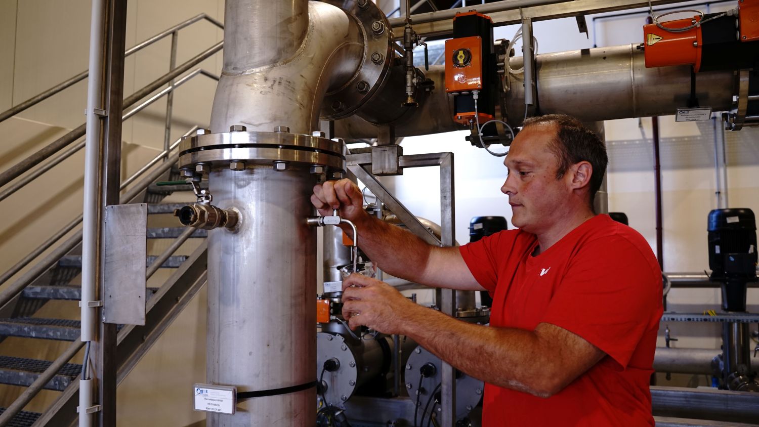 Mann in rotem T-Shirt bedient Anlage mit Edelstahlrohren und Messbecher in Industriehalle
